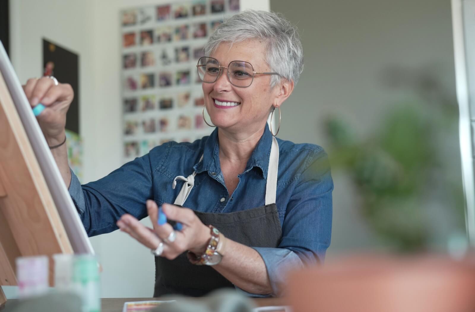 A smiling senior woman with paints on a canvas, wearing a denim shirt and apron in a bright, creative space. Show less