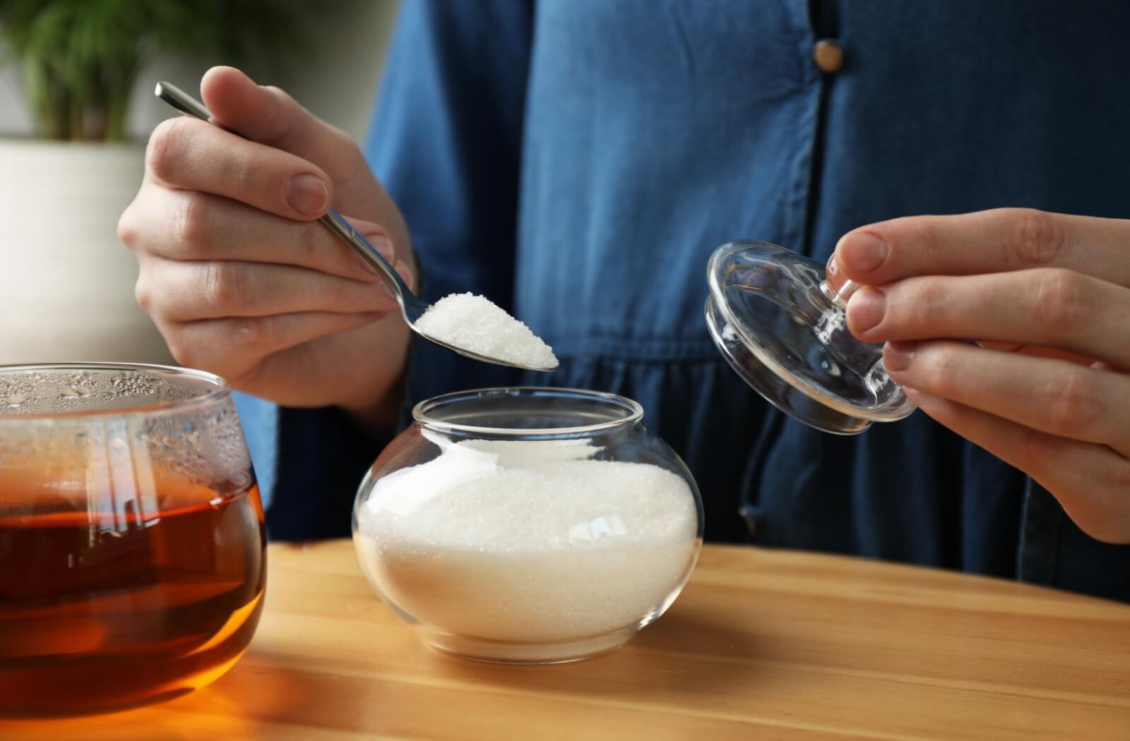 An older adult taking a spoonful of artificial sweetener to mix into a clear cup of iced tea.
