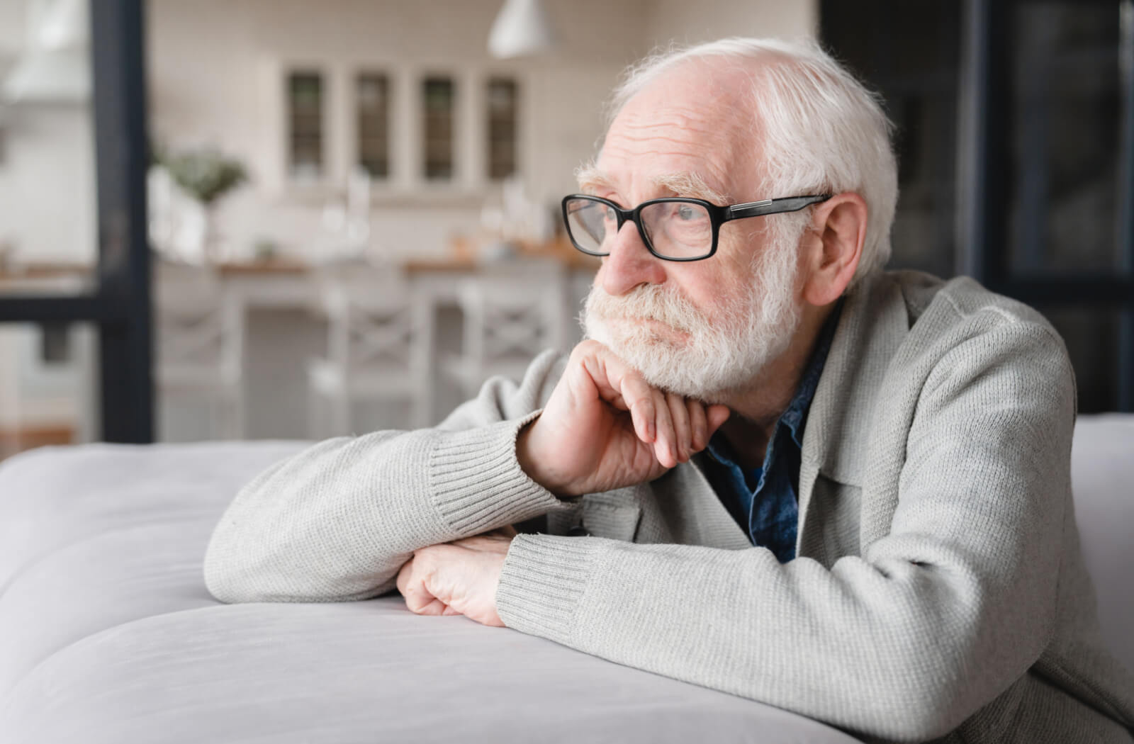 An older adult with glasses on sitting on a couch and looking out the window with a serious expression