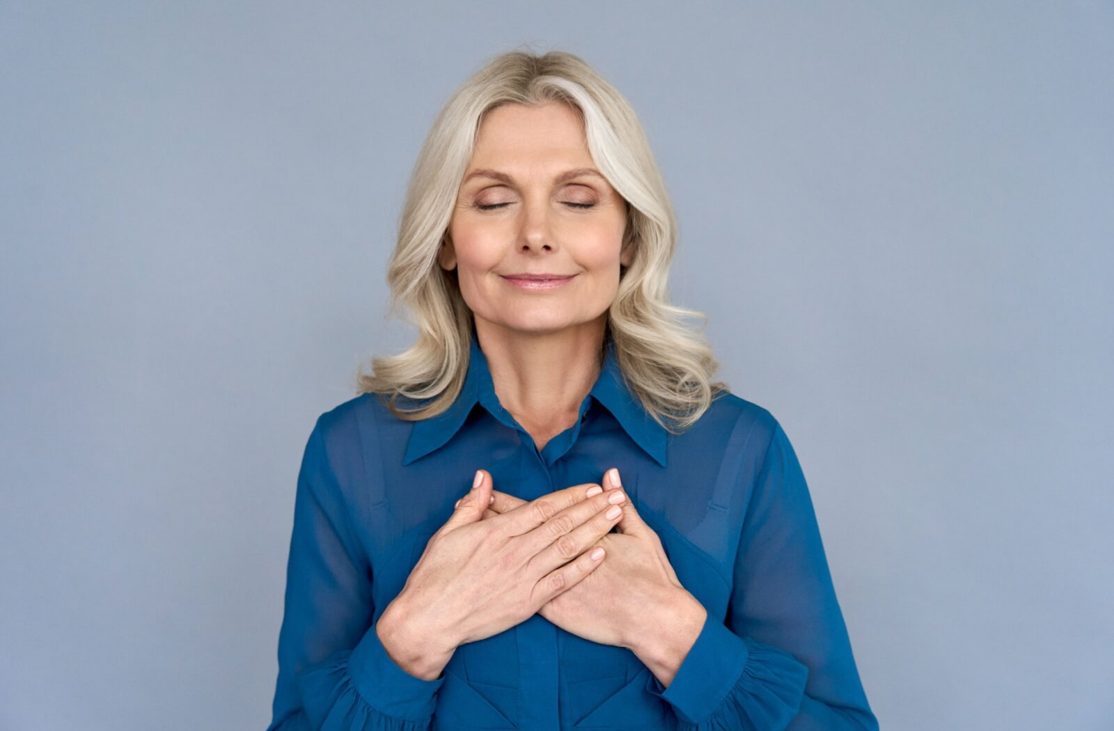 Against a grey-blue background, an older adult in a blue shirt closes their eyes and places both hands over their heart